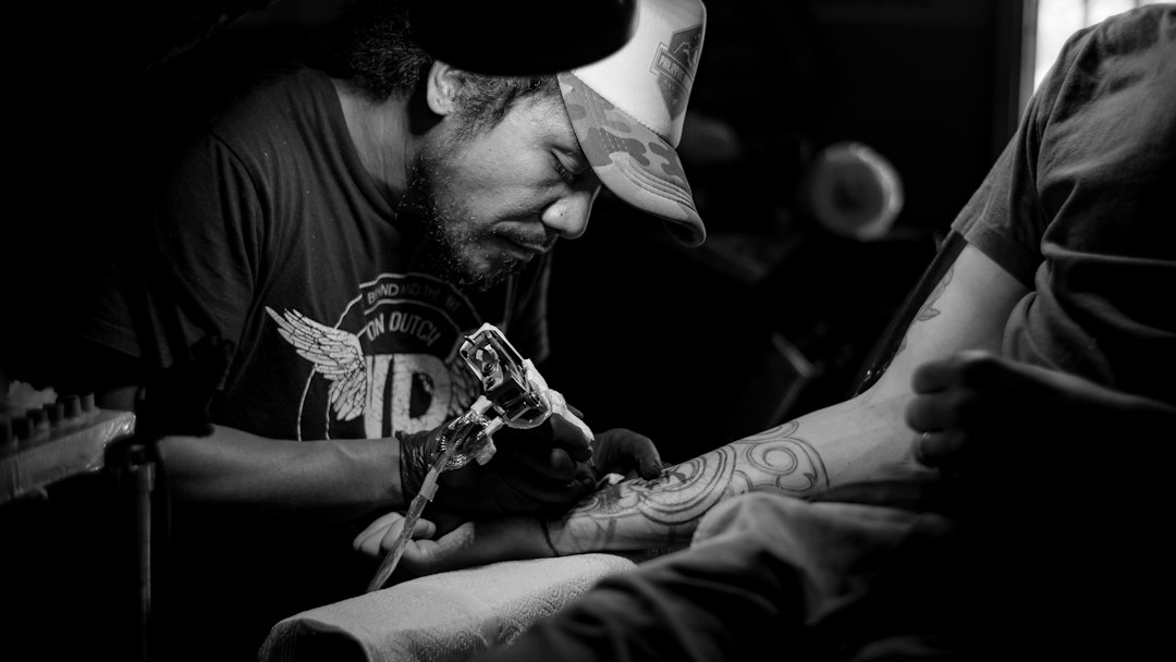 Tatuator w czapce tatuuje ramię klienta, czarno-białe